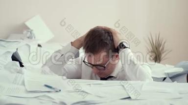 一个戴眼镜的疲惫的年轻人坐在一堆文件和签名文件里。 因为疲劳，一个人心烦意乱